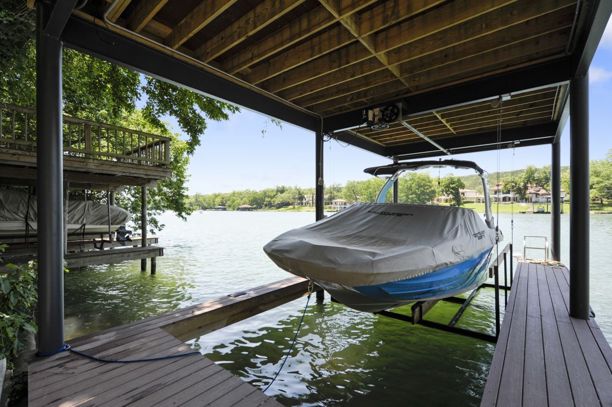 A covered private boat dock and mechanical lift housing a blue wakeboarding boat on the calm waters of Lake Austin, showcasing the premier waterfront amenities available to guests staying with Walker Luxury Vacation Rentals.