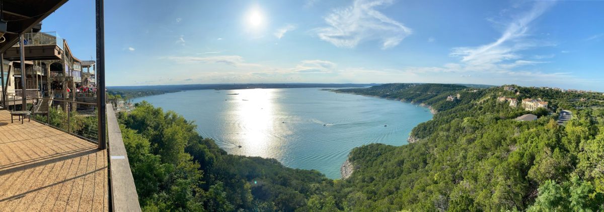 A breathtaking panoramic view of Lake Travis from the balcony of The Oasis restaurant in Austin, Texas. The sun reflects off the sparkling blue water surrounded by lush green hills, showcasing the iconic scenery near Walker Luxury Vacation Rentals' hill country properties.