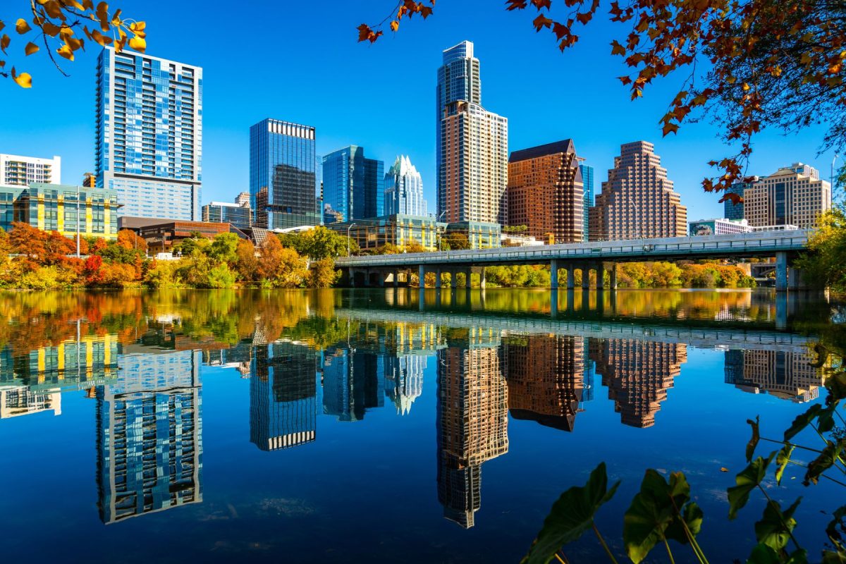 A stunning autumn view of the Austin, Texas downtown skyline perfectly reflected in the calm waters of Lady Bird Lake. Vibrant orange and yellow fall foliage frames the modern skyscrapers under a clear blue sky, highlighting the scenic urban beauty near Walker Luxury Vacation Rentals' properties.